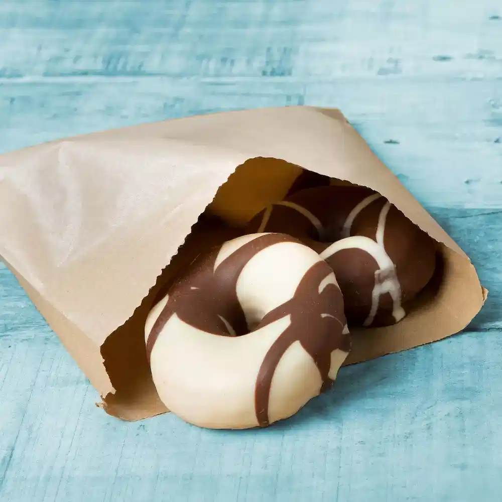Pączki w czekoladzie częściowo widoczne w brązowej papierowej torbie na niebieskiej drewnianej powierzchni.