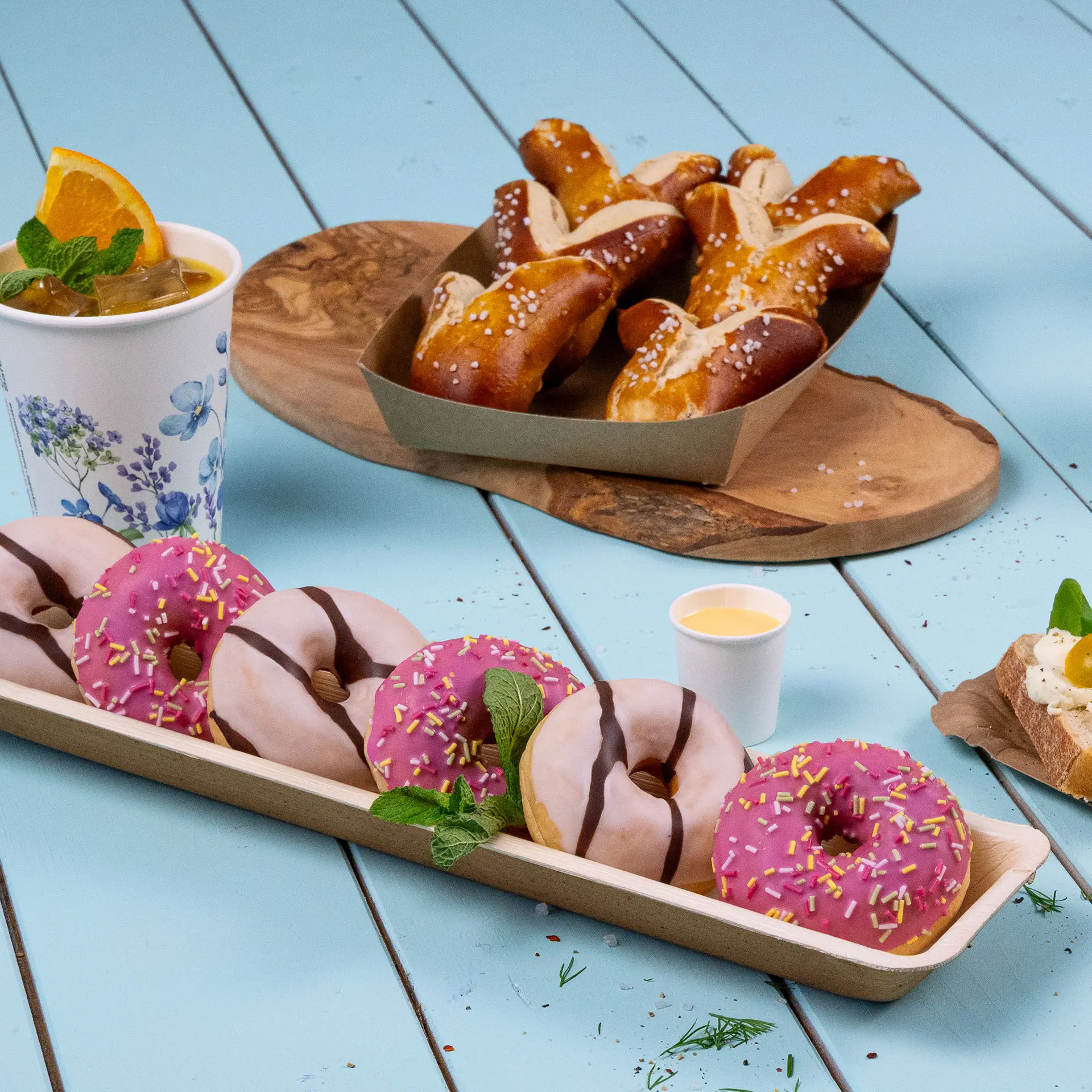 Taca z różowymi i białymi donutami z polewą czekoladową, kubkiem napoju i preclami na jasnoniebieskiej powierzchni.