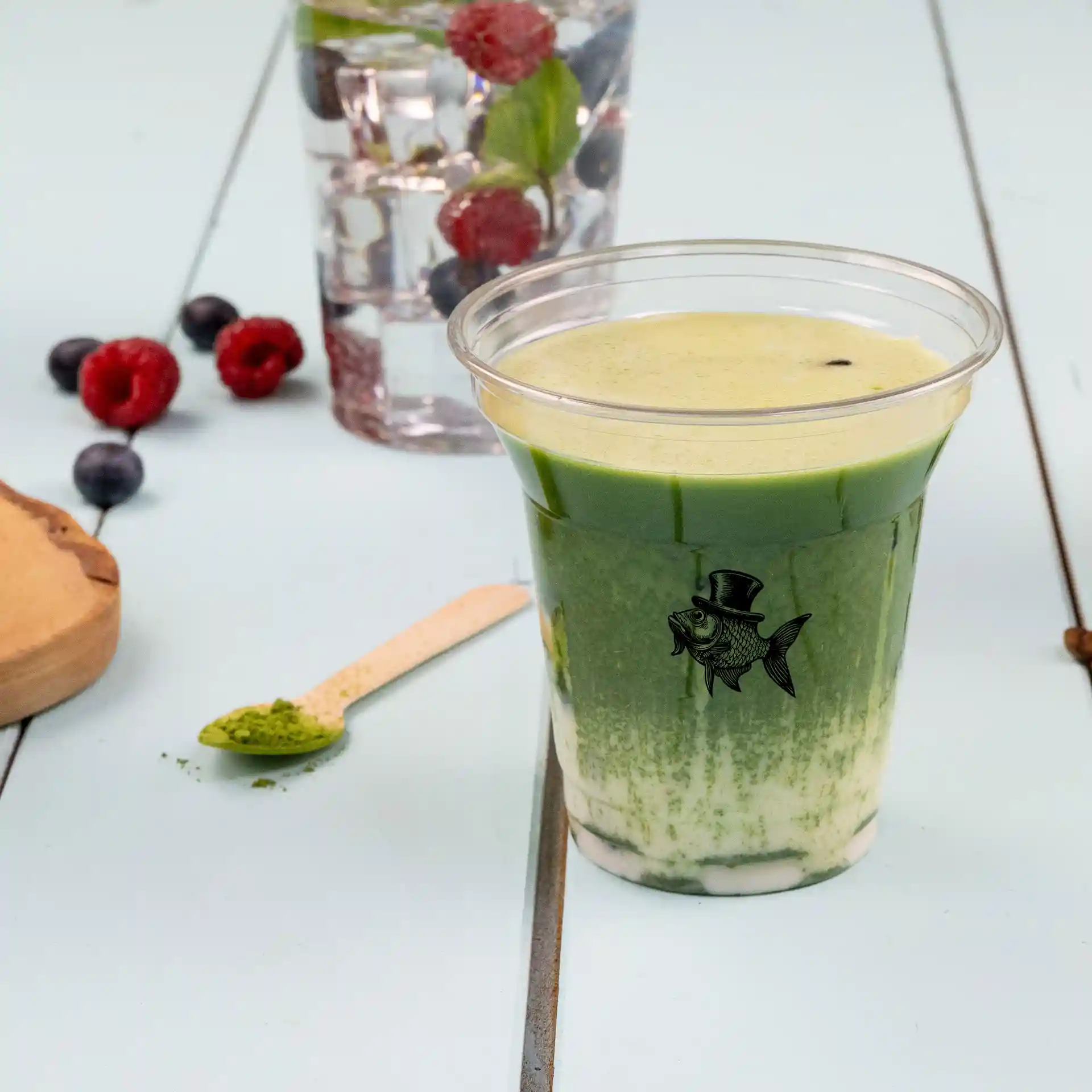 Przezroczysty kubek rPET 8 oz wypełniony zielonym napojem, z logo ryby, drewnianą łyżką i jagodami w tle.