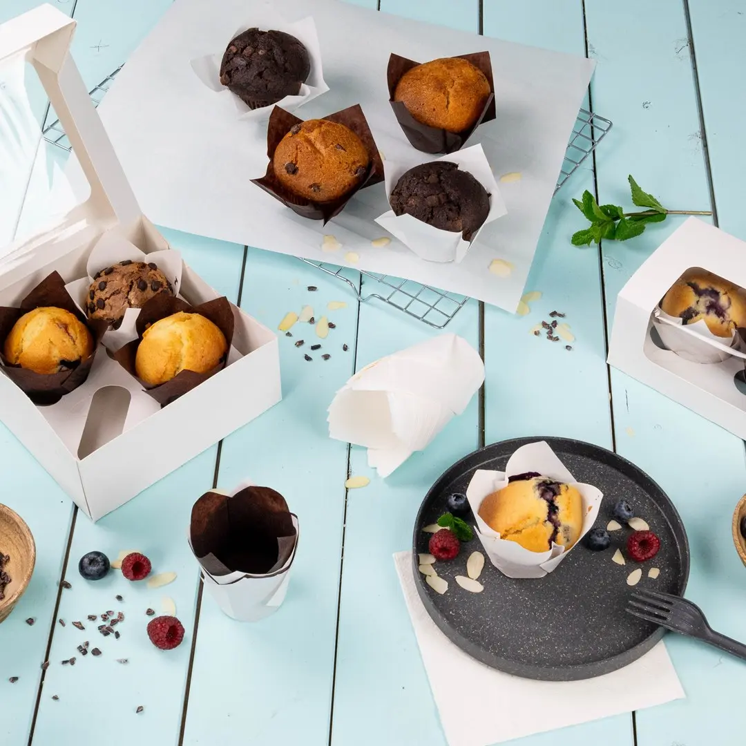 Różne muffiny w papierowych opakowaniach na jasnoniebieskiej powierzchni, z białym pudełkiem i talerzem muffinek oraz jagód.