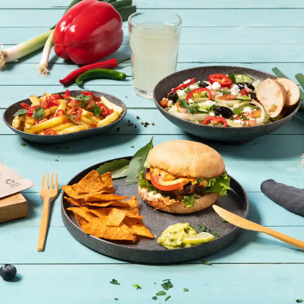 Burger z sałatą, pomidorem i sosem, podawany z chipsami tortilla i guacamole, obok sałatki i frytek.