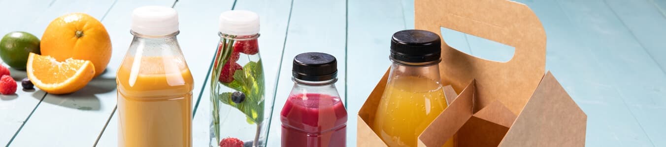 Cztery butelki napojów na jasnoniebieskiej powierzchni, z różnymi owocami, w tym pomarańczą i limonką. Papierowy nosidło trzyma dwie butelki.