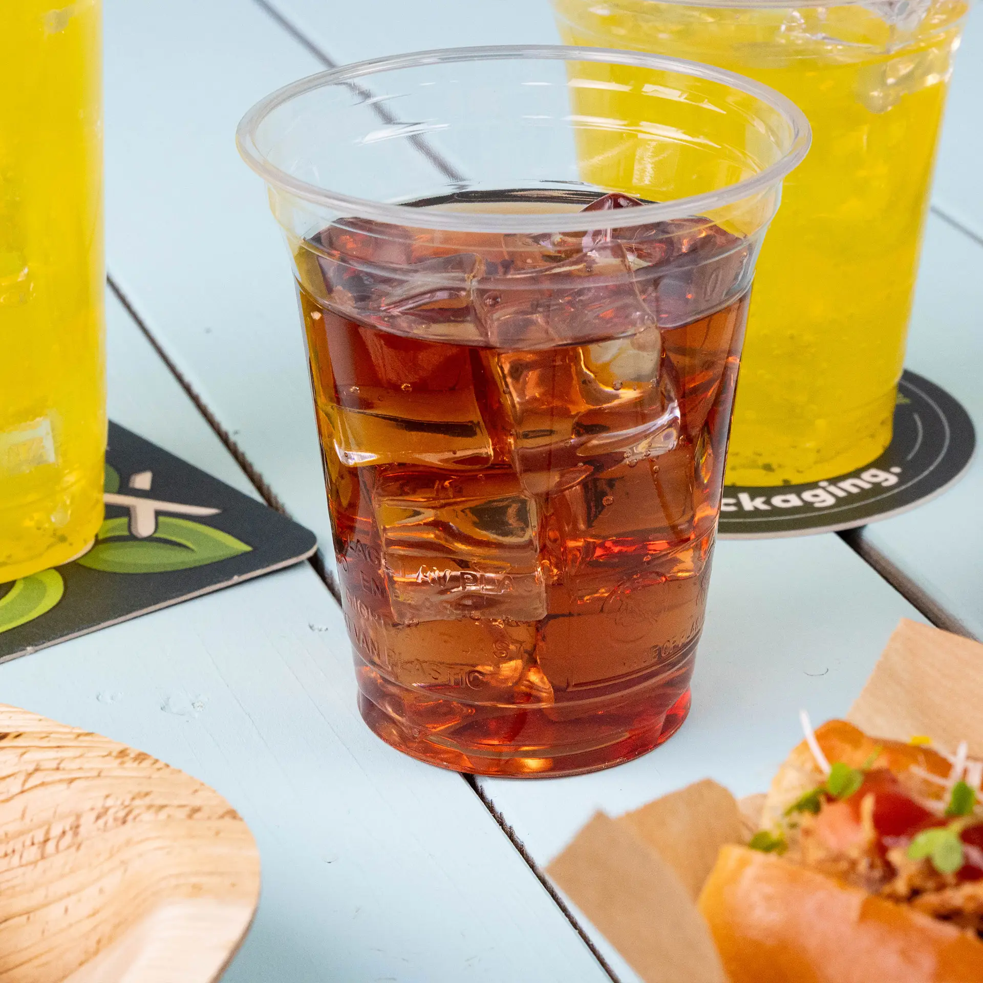 Przezroczysty plastikowy kubek 12 oz z bioplastiku PLA wypełniony mrożoną herbatą, otoczony żółtymi napojami i jedzeniem.